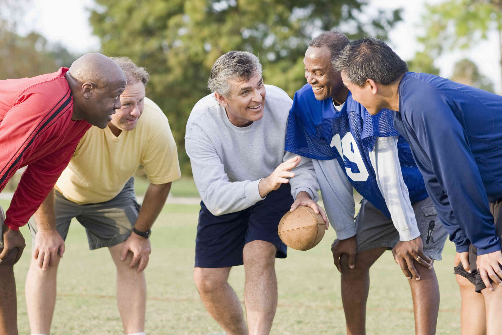 A group of men smile and chat as they crouch around in a huddle outside. Some of them wear mesh, colored vests and one of them holds a football.